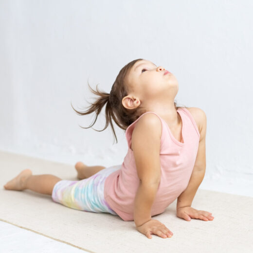 Young child in a pink shirt and colorful pants practices a yoga pose on a mat indoors, arching her back and looking upward.