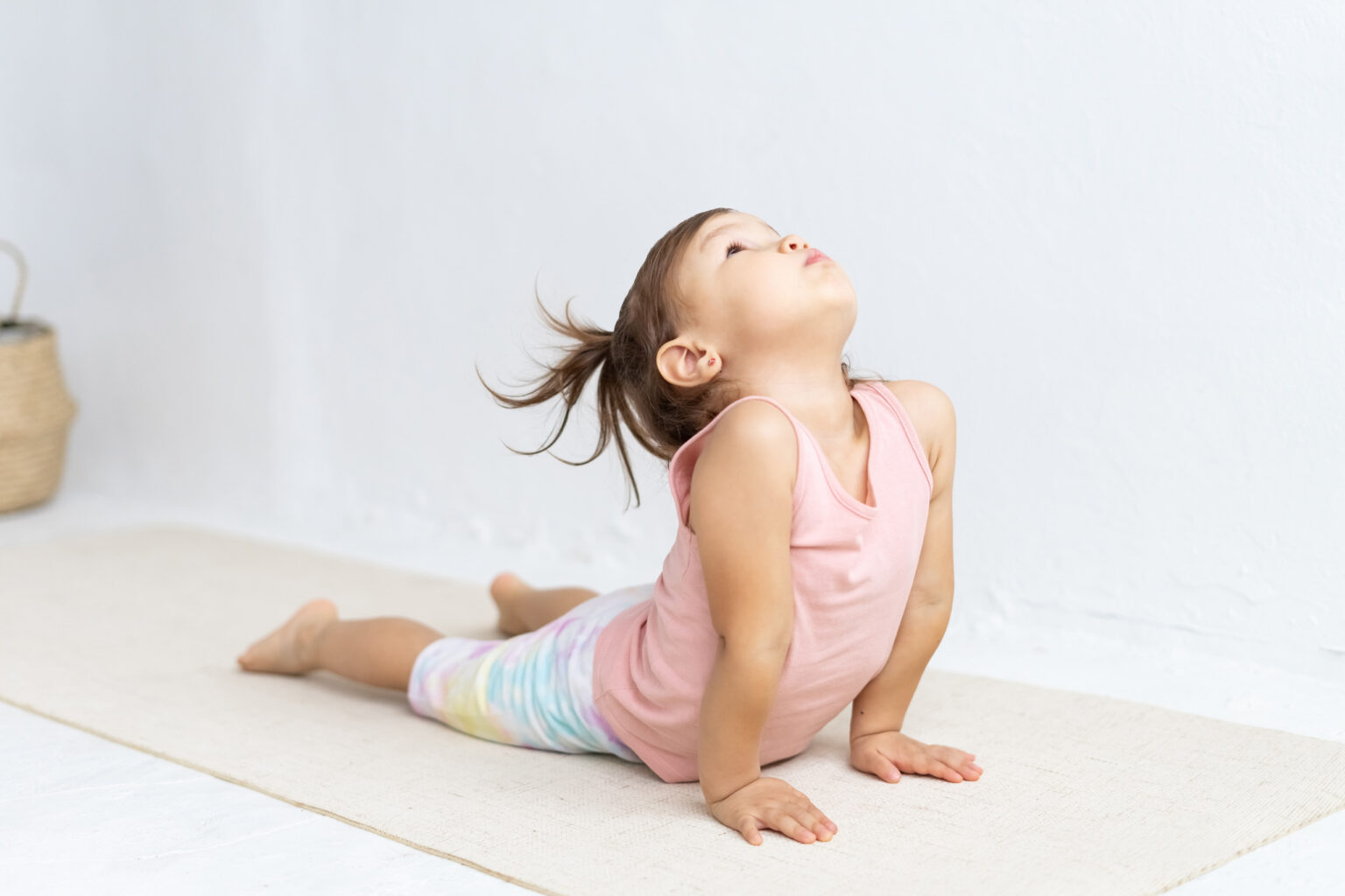 Young child in a pink shirt and colorful pants practices a yoga pose on a mat indoors, arching her back and looking upward.