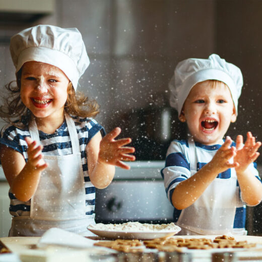 Two young children in chef hats and aprons laugh and clap in a kitchen, causing flour to fly through the air around them.