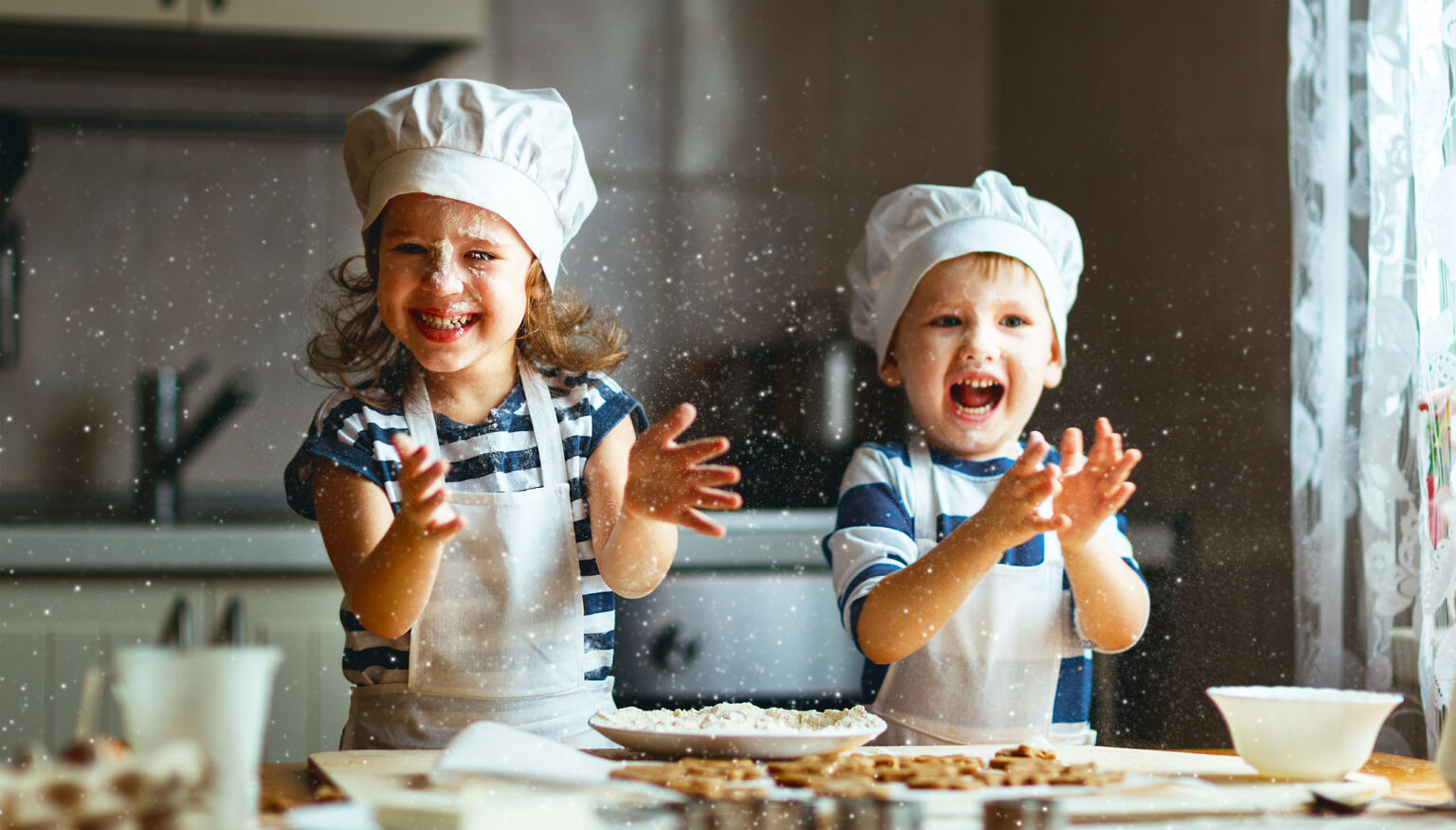 Two young children in chef hats and aprons laugh and clap in a kitchen, causing flour to fly through the air around them.
