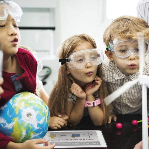Four children wearing safety goggles observe a small model wind turbine in a classroom, with a globe and educational materials on the table. An adult stands nearby.