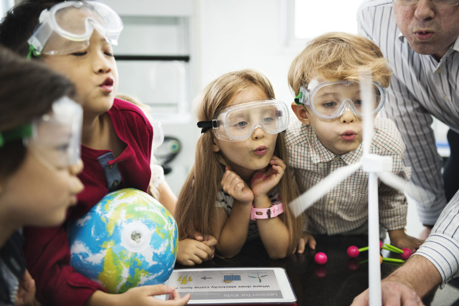 Four children wearing safety goggles observe a small model wind turbine in a classroom, with a globe and educational materials on the table. An adult stands nearby.