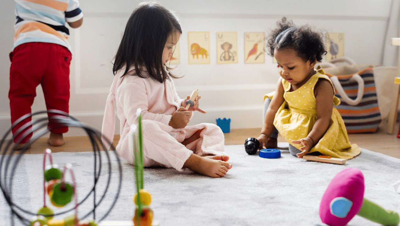 Three young children play on a carpeted floor with various toys in a brightly lit room decorated with animal pictures on the wall.