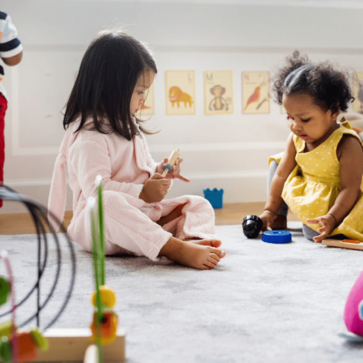 Three young children play on a carpeted floor with various toys in a brightly lit room decorated with animal pictures on the wall.