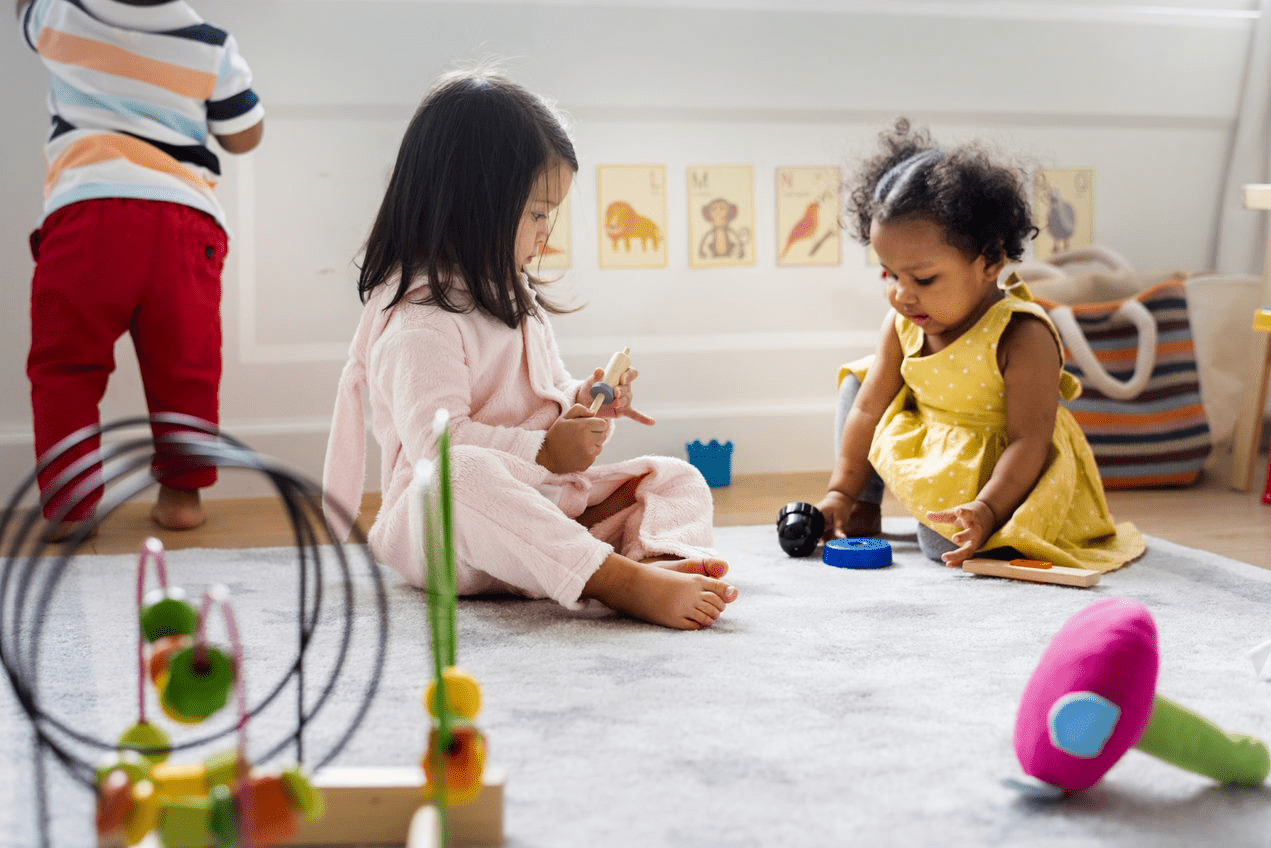 Three young children play on a carpeted floor with various toys in a brightly lit room decorated with animal pictures on the wall.