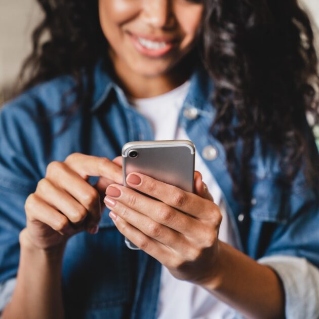 A person with long curly hair, wearing a denim shirt, is smiling and using a smartphone while sitting indoors.