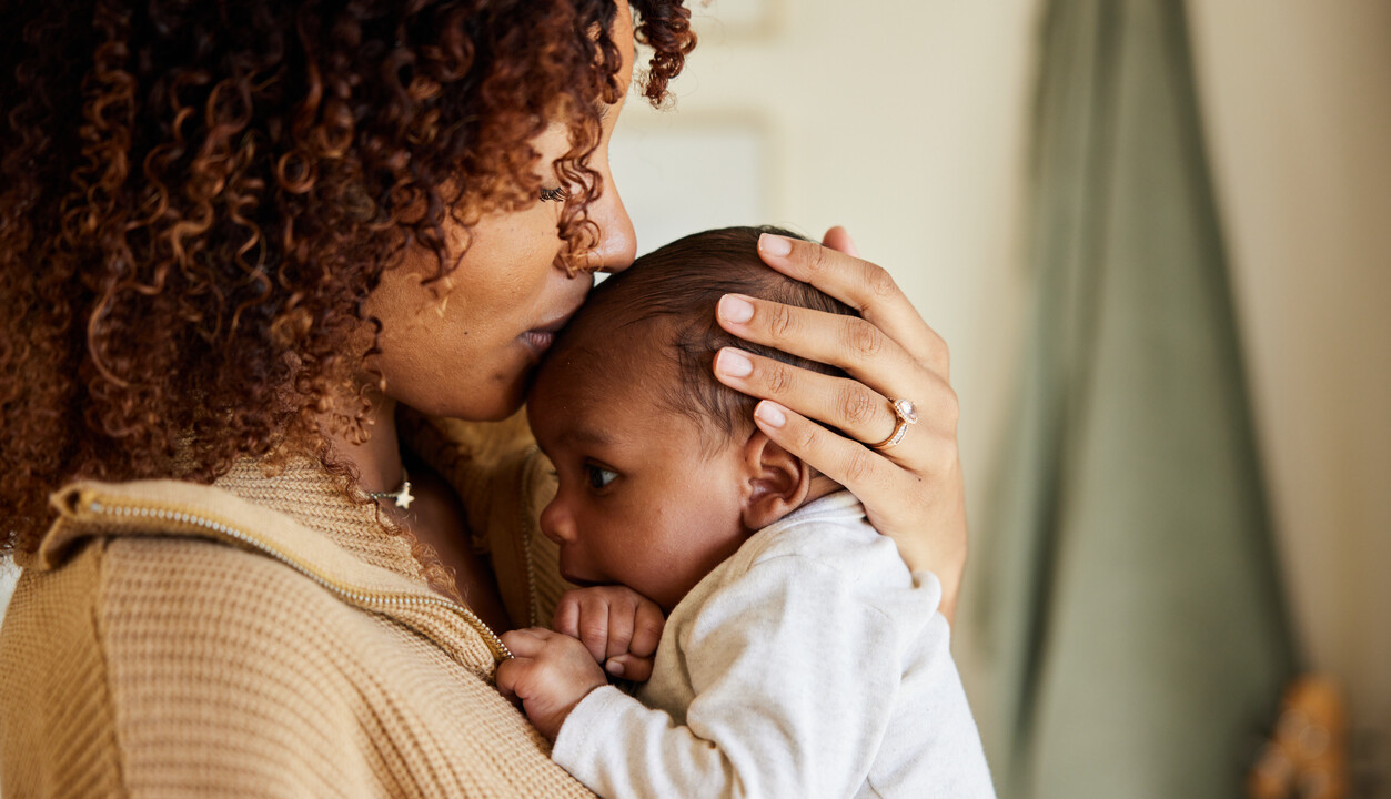 A woman holds a baby close to her chest and kisses the baby’s head. Both are indoors, and the woman wears a beige sweater.