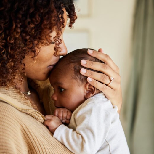 A woman holds a baby close to her chest and kisses the baby’s head. Both are indoors, and the woman wears a beige sweater.