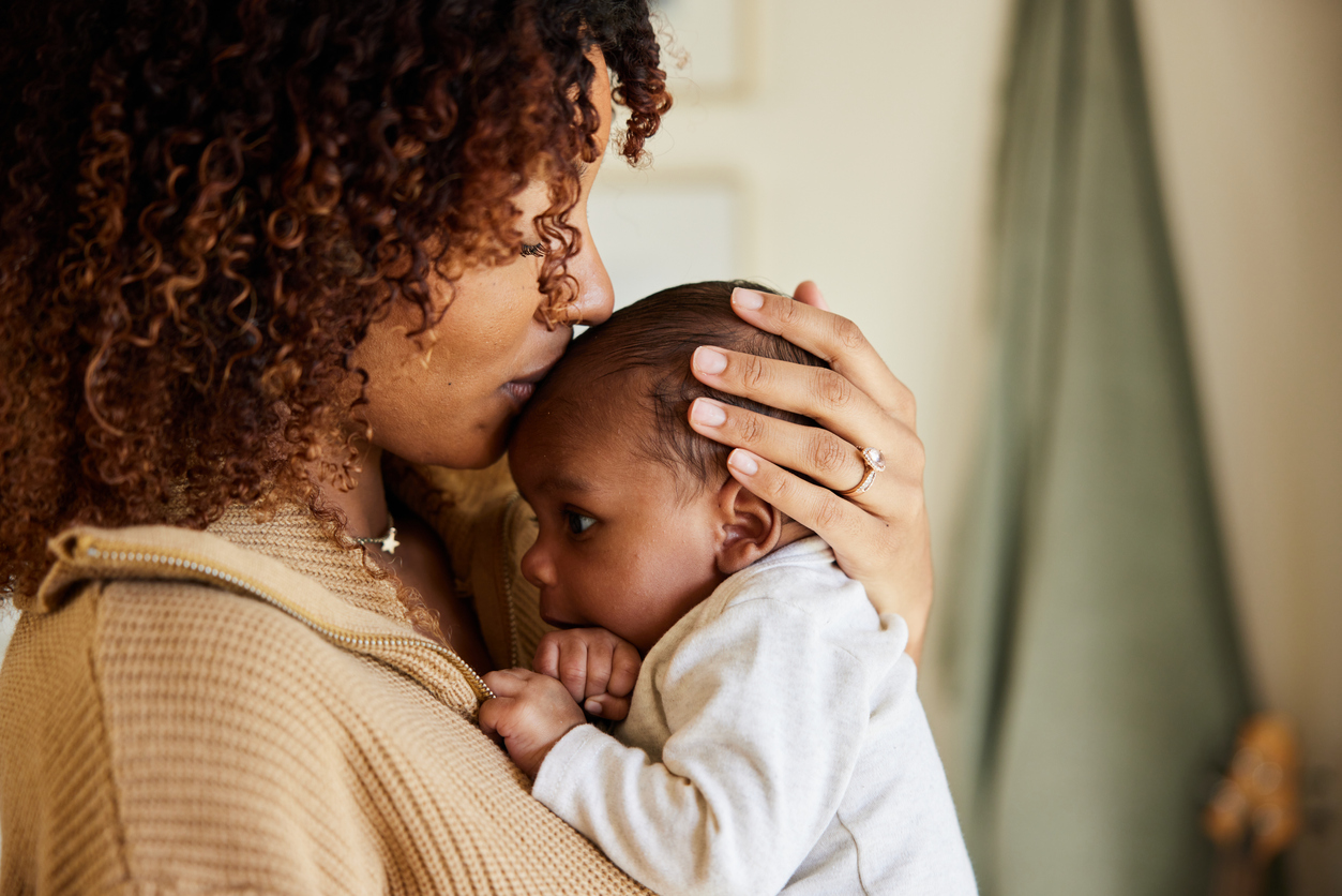 A woman holds a baby close to her chest and kisses the baby’s head. Both are indoors, and the woman wears a beige sweater.