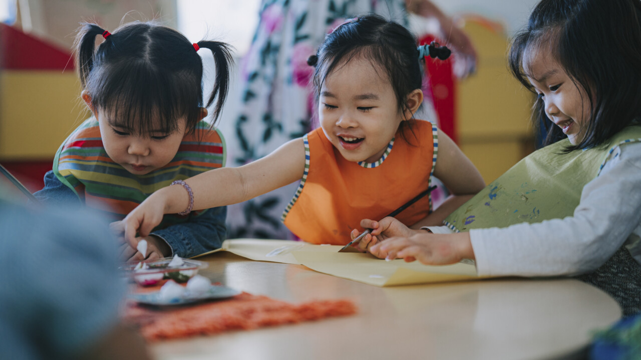 Three young children sit at a round table, drawing and painting together with art supplies, while an adult stands in the background.