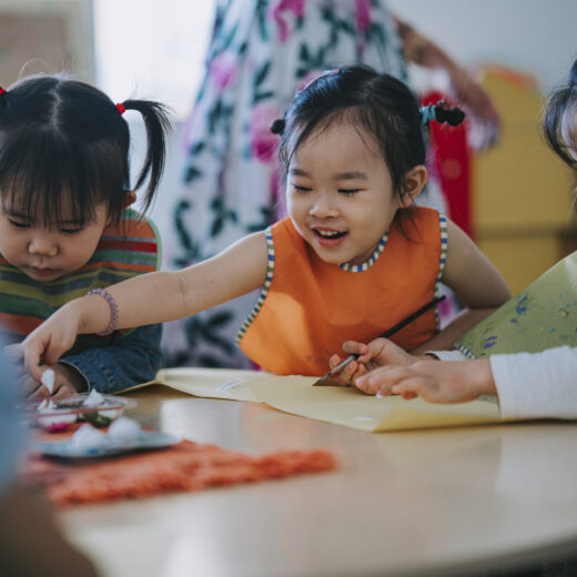 Three young children sit at a round table, drawing and painting together with art supplies, while an adult stands in the background.