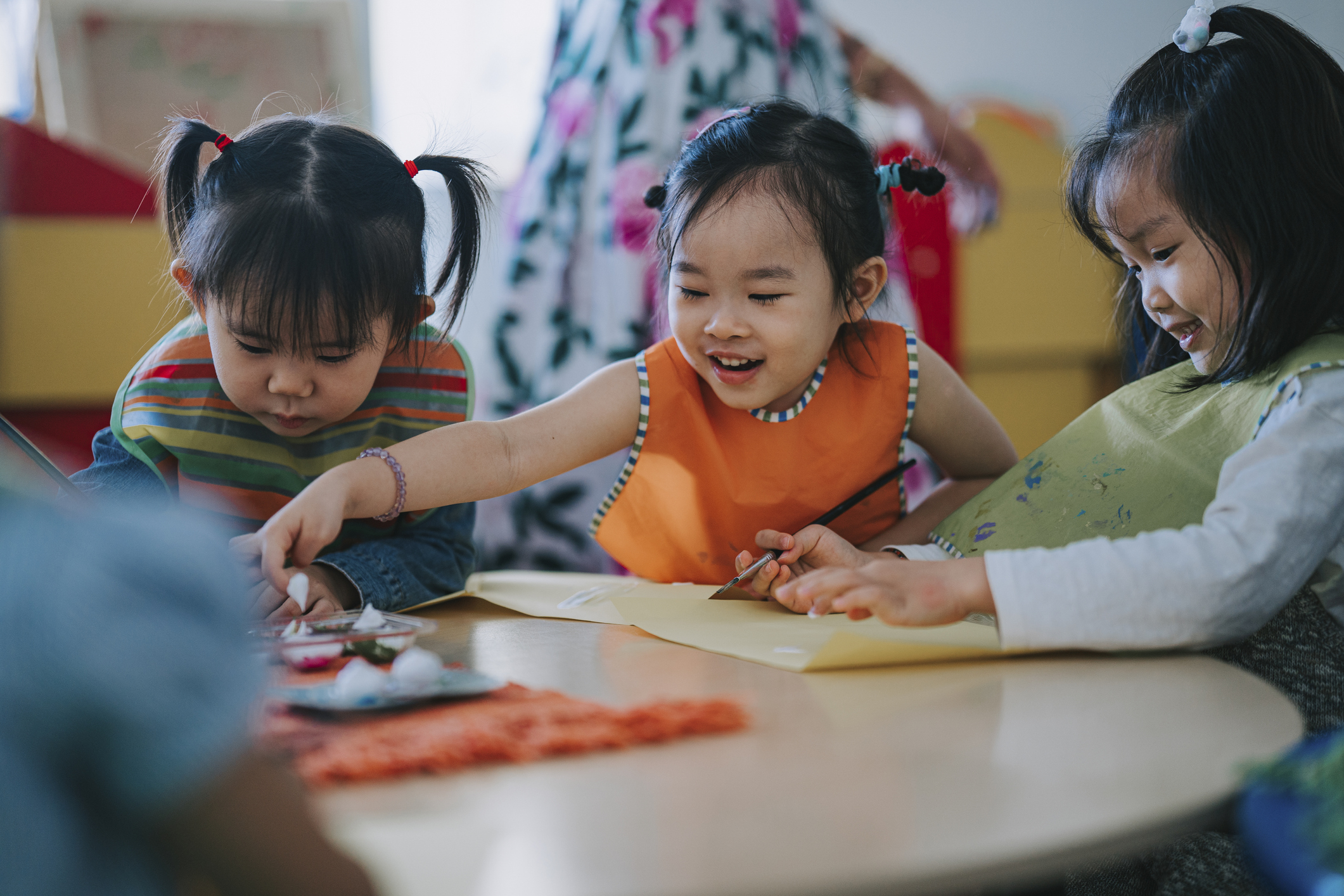 Three young children sit at a round table, drawing and painting together with art supplies, while an adult stands in the background.