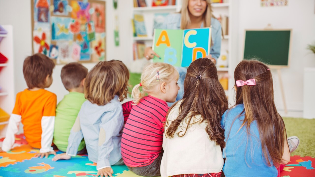 A teacher shows alphabet cards to a group of young children sitting on a colorful alphabet mat in a classroom.