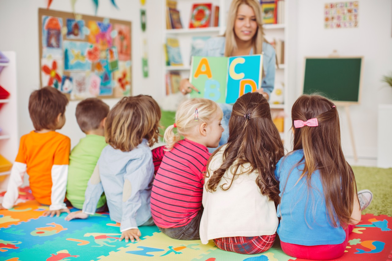 A teacher shows alphabet cards to a group of young children sitting on a colorful alphabet mat in a classroom.