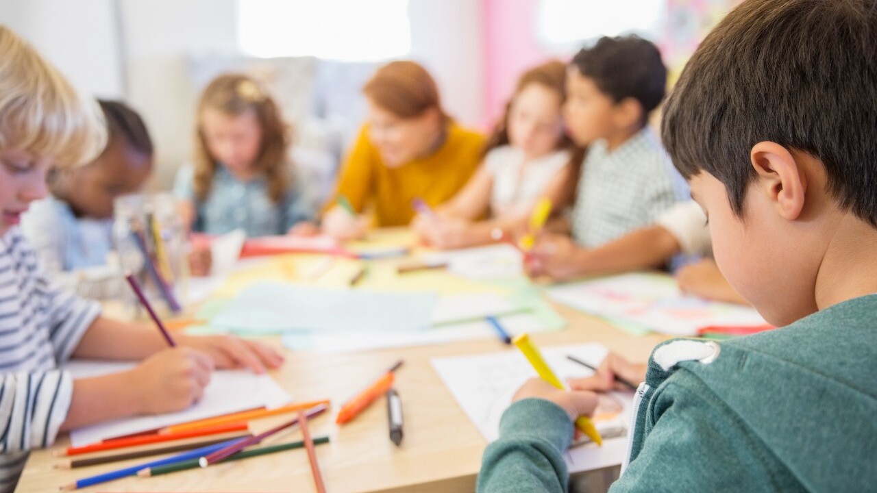 Children sit around a table drawing with colored pencils and paper, with a teacher supervising in a classroom setting.
