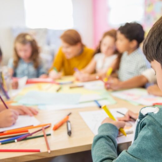 Children sit around a table drawing with colored pencils and paper, with a teacher supervising in a classroom setting.
