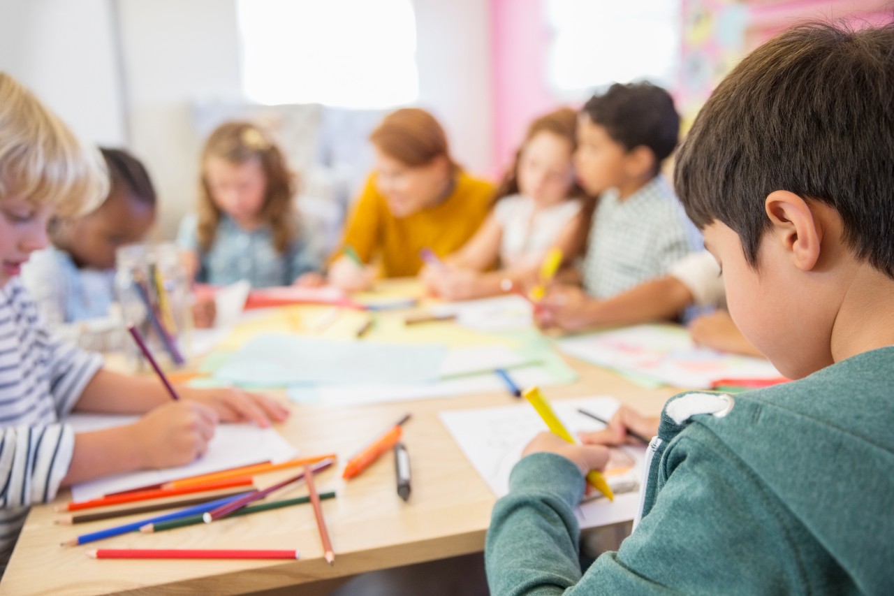 Children sit around a table drawing with colored pencils and paper, with a teacher supervising in a classroom setting.
