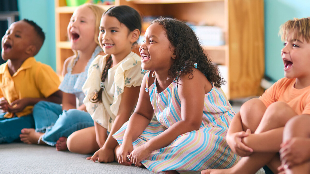 A group of young children sit on the floor in a classroom, smiling and laughing together.