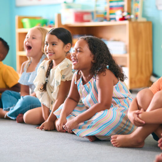 A group of young children sit on the floor in a classroom, smiling and laughing together.