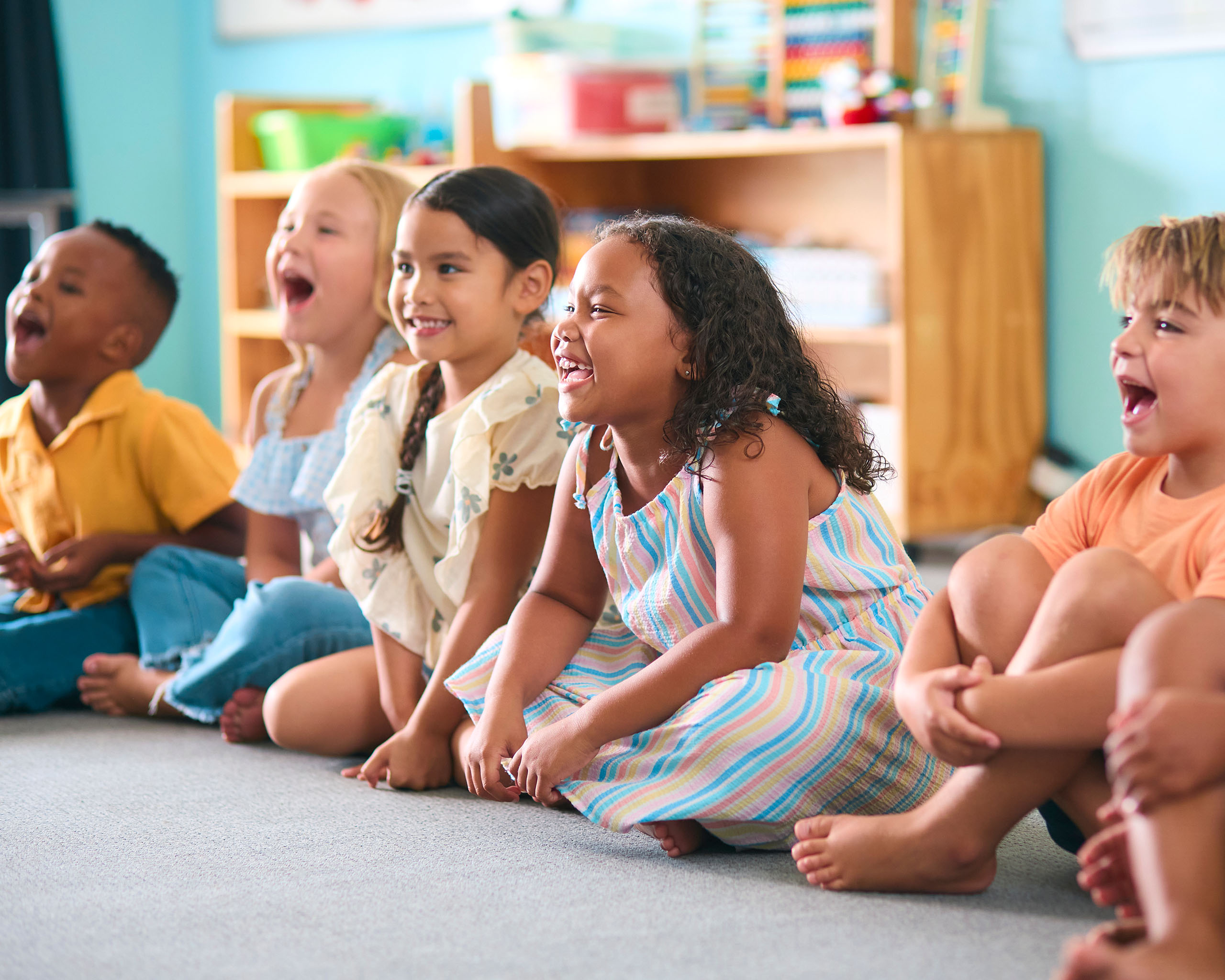 A group of young children sit on the floor in a classroom, smiling and laughing together.