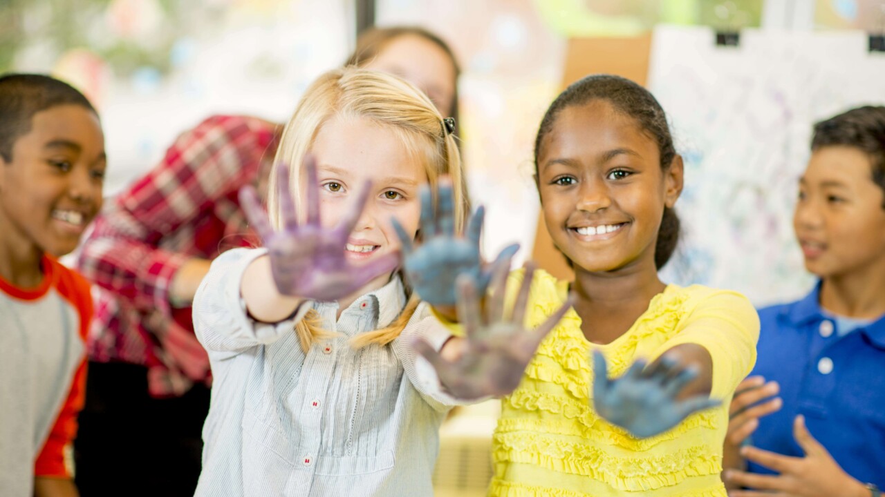 Two children stand in front, smiling and showing their paint-covered hands, with two other children visible in the background in a classroom setting.