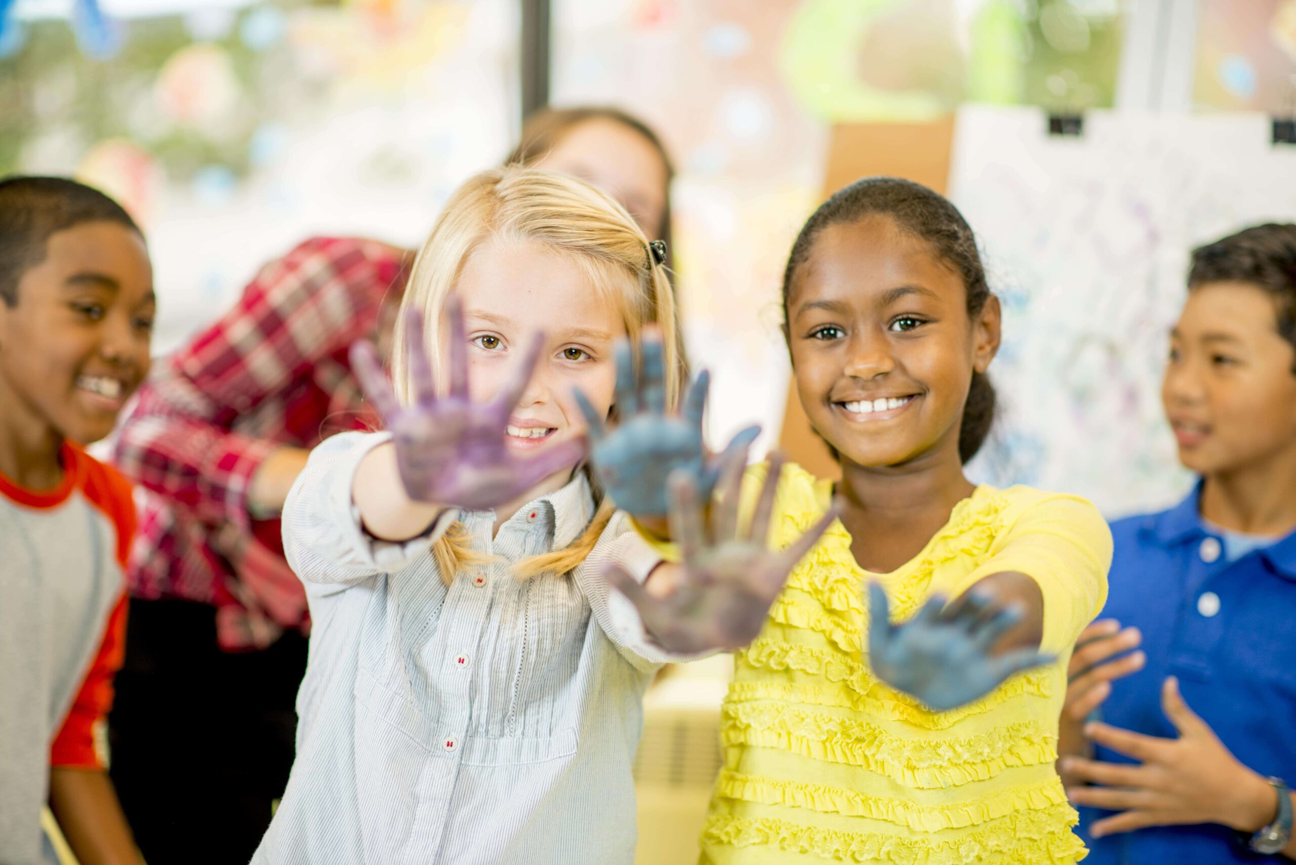 Two children stand in front, smiling and showing their paint-covered hands, with two other children visible in the background in a classroom setting.