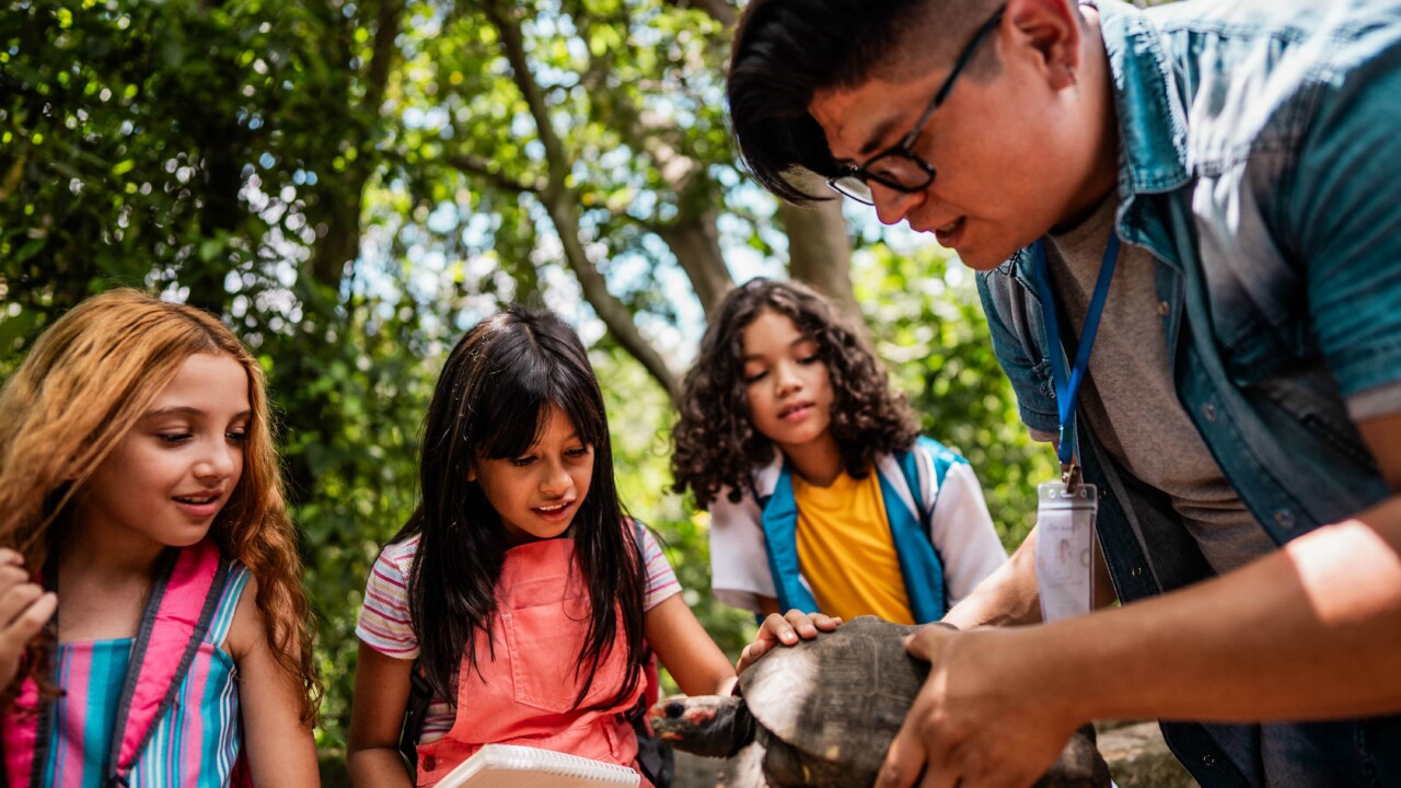 A group of children observe a person holding a turtle outdoors, with one child taking notes and trees in the background.