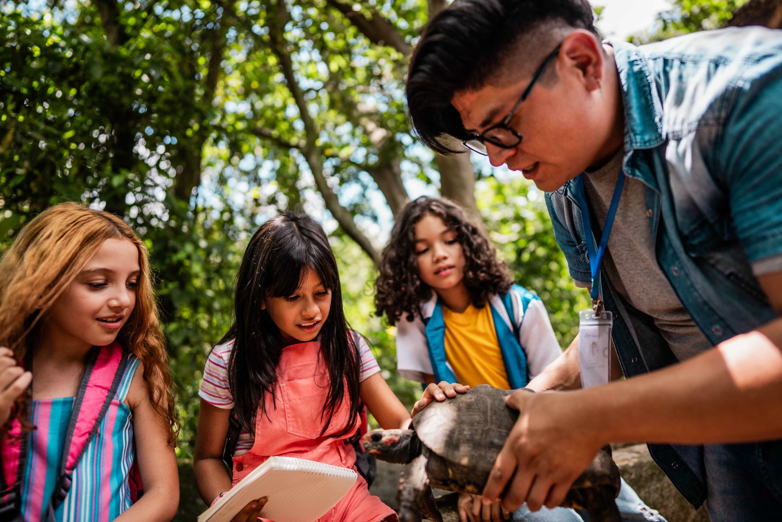 A group of children observe a person holding a turtle outdoors, with one child taking notes and trees in the background.
