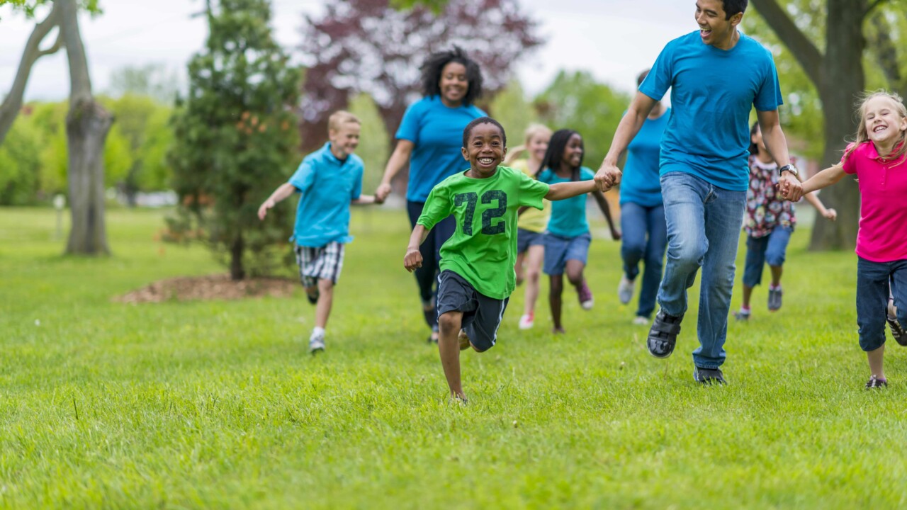 A group of children and adults run together outdoors on a grassy field, holding hands and smiling. Trees are visible in the background.