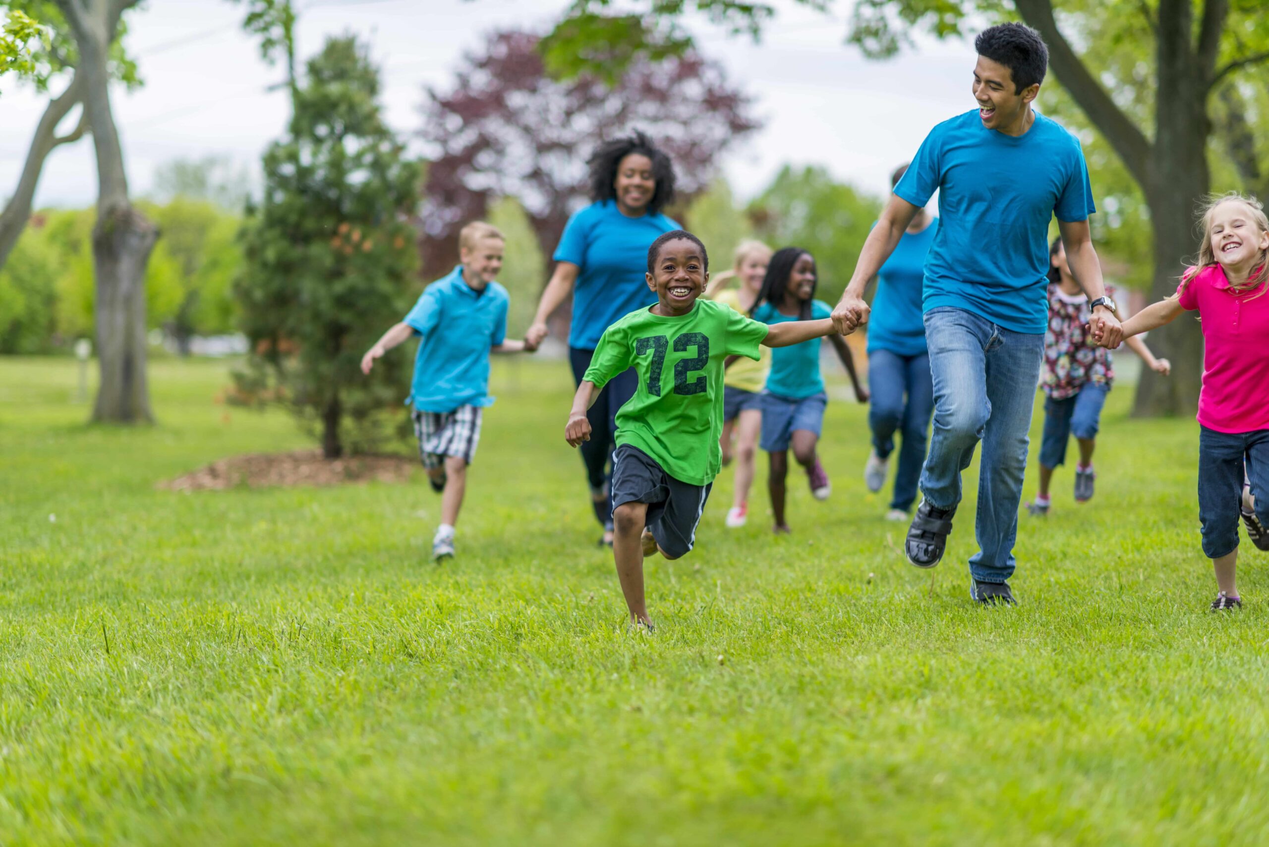 A group of children and adults run together outdoors on a grassy field, holding hands and smiling. Trees are visible in the background.